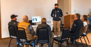 Man standing in front of a computer presenting to a seated group of five people.